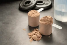 Two scoops of protein powder on a countertop with weight plates and a shaker bottle in the background, illustrating common supplements used to support muscle repair and recovery in a blog post about eating enough protein titled "Are you Eating enough protein?".