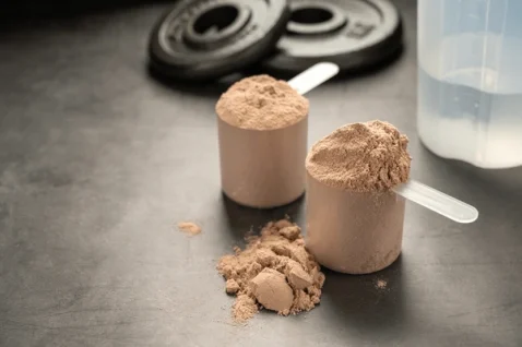Two scoops of protein powder on a countertop with weight plates and a shaker bottle in the background, illustrating common supplements used to support muscle repair and recovery in a blog post about eating enough protein titled "Are you Eating enough protein?".